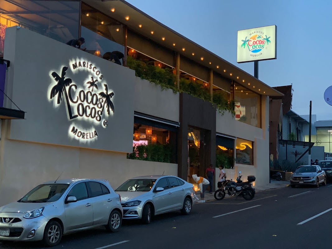 Patmos Cocina de Mar – exterior view of the restaurant at night, lights on, with a waiter carrying a plate of grilled octopus