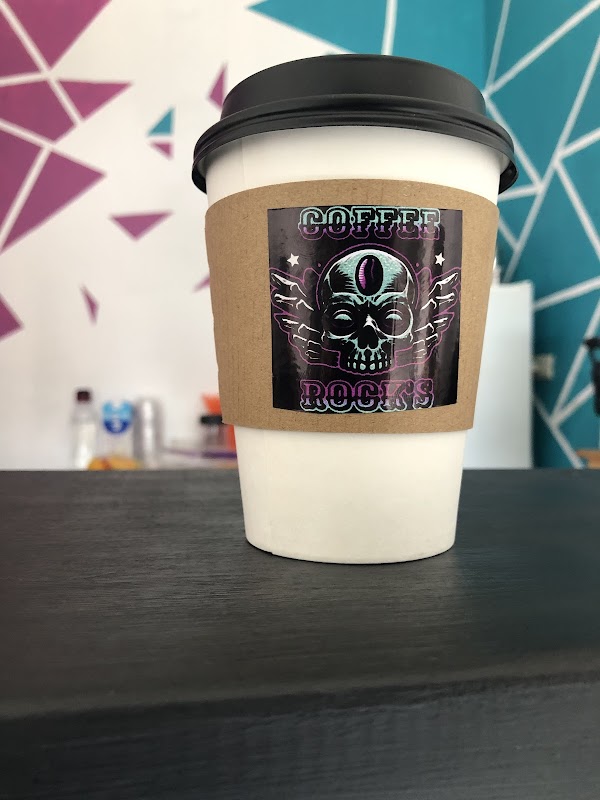 Interior de Coffee Rock’s con barra de espresso, taza humeante y clientes en mesas de madera