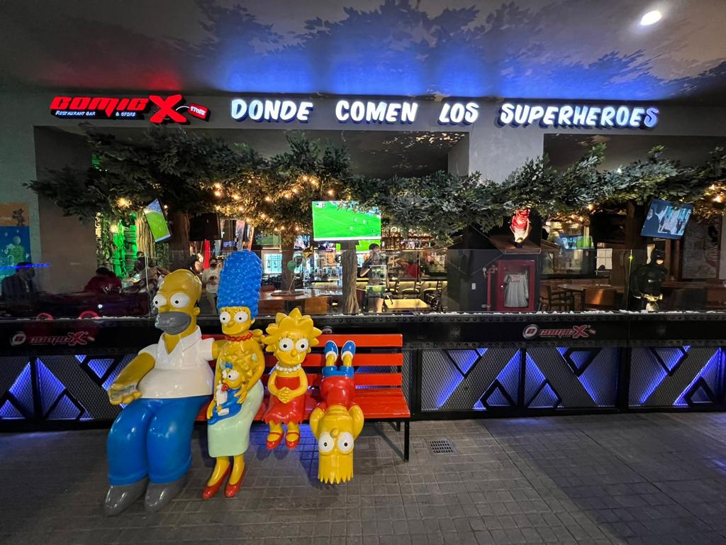 Comicx Otay exterior with superhero signage and neon lights, showing the bar entrance and a patron holding a themed cocktail